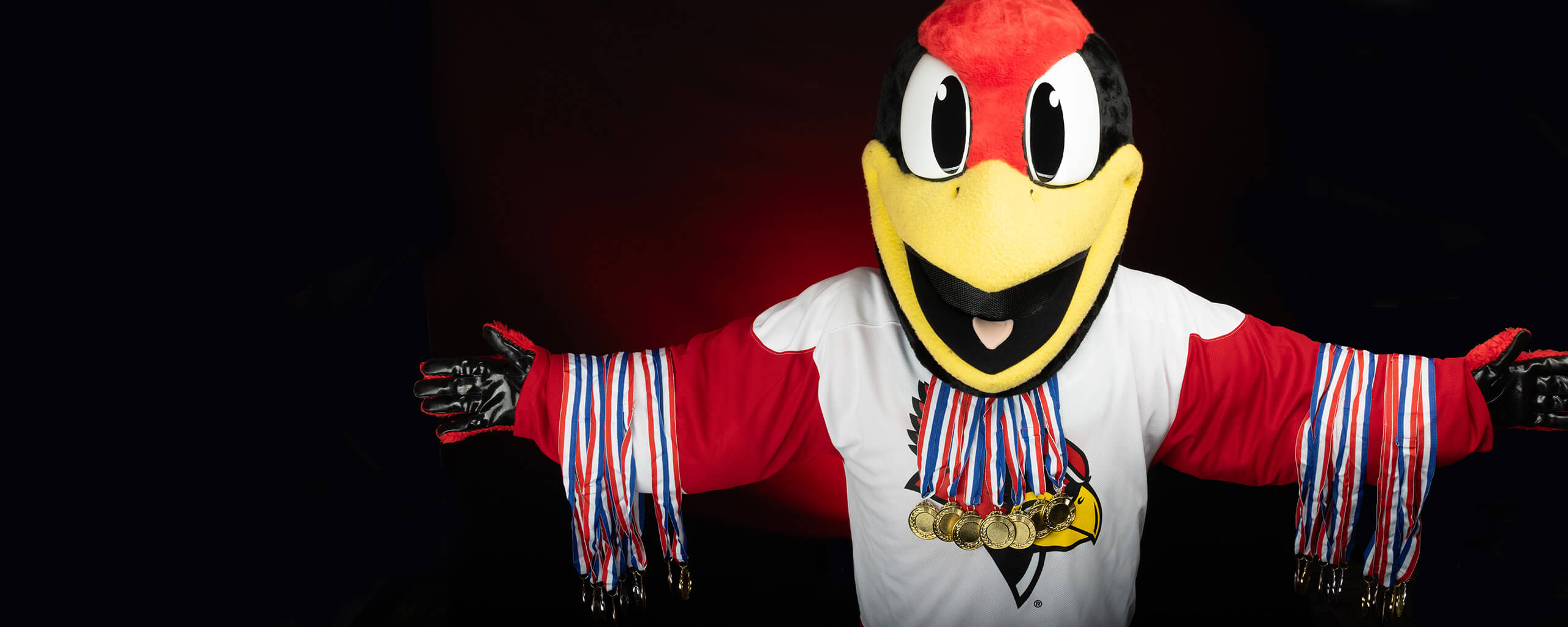 Reggie Redbird, arms spread outward, wears multiple gold medals around his neck and arms.