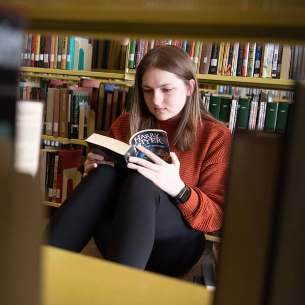 A student sits between the aisles of the library, reading a book.